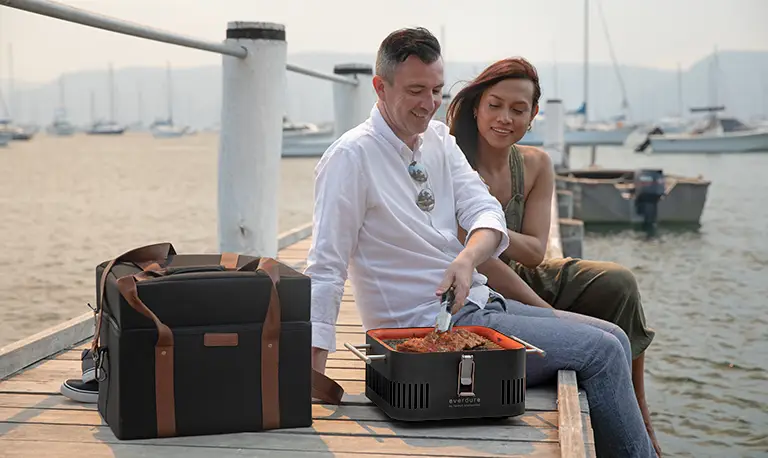 couple cooking on an everdure cube bbq while sat on a jetty by the sea
